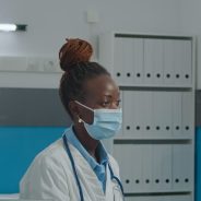 Medical team of specialists consulting senior patient in cabinet while wearing face masks for virus protection. Doctor and nurse talking to elder person behind plexiglass wall about treatment